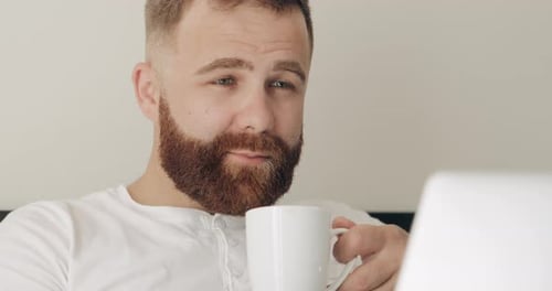 Close Up View of Concentrated Man Drinking Hot Coffee with Rich Aroma While Working in Bed, Handsome