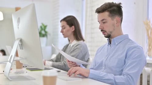 Homme créatif sérieux faisant de la paperasse et utilisant un ordinateur de bureau