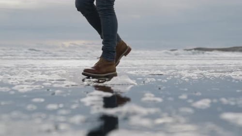 Man In Hiking Boots Walking Along Wet Beach And Through Surf