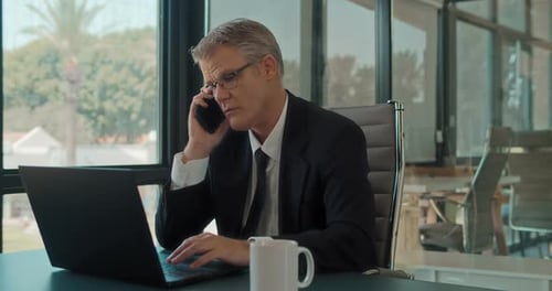 A businessman talking on the phone while working on a laptop at the office.