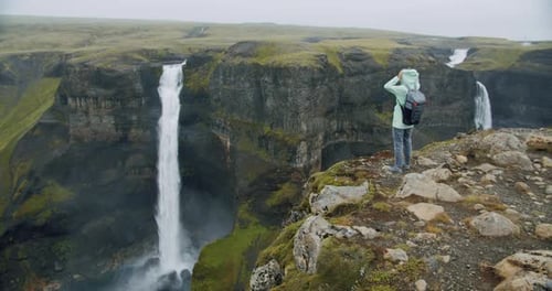 Frau mit Rucksack setzt die Outdoor-Mantelhaube vor dem Haifoss-Wasserfall im isländischen Hochland auf
