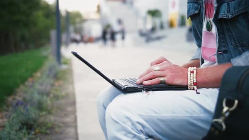 Close Up Shot of Young Hipster Man and in Hat Sits on Bench in City Center in Park Using Laptop Slow