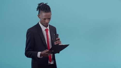 Black Man in an Office Suit and with Dreadlocks Reads the Text on a Paper Tablet and Holding Glass