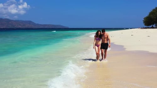 Young couple in swimsuit walking around exotic beach over white sand washed by waves of blue turquoi