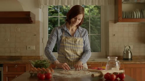 30s Woman in Apron Placing Grated Cheese on Pizza Dough Standing in the Kitchen