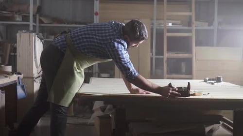 Professional Male Cabinetmaker in Shirt Planing Wood in a Dark Authentic Workshop