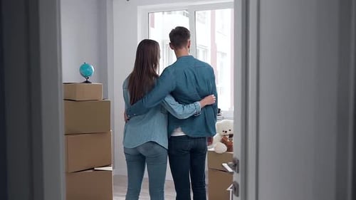 Happy Couple which Standing Embracing in the Middle of the Empty New flat in Front of Window