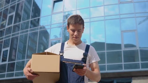 A Delivery Worker Holds a Box in His Hands and Talks on a Cell Phone