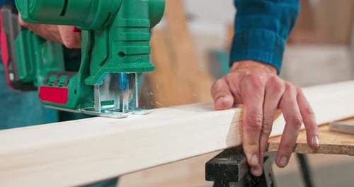 Young Carpenter Cuts a Beam with a Jigsaw Craftsman's Hands in Cloth Protective Gloves The Jigsaw