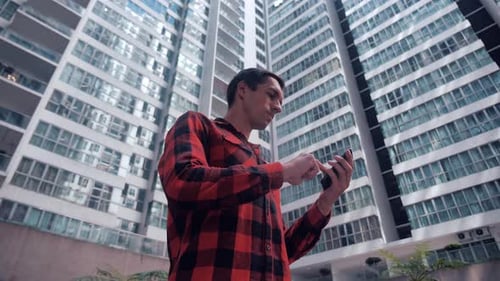 Handsome Young Man in Plaid Shirt Browsing His Smartphone in City on Urban Buildings Background