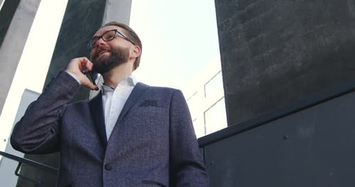 Man in Glasses which Standing Outdoors on the Steps of Office Center and Has Mobile Conversation