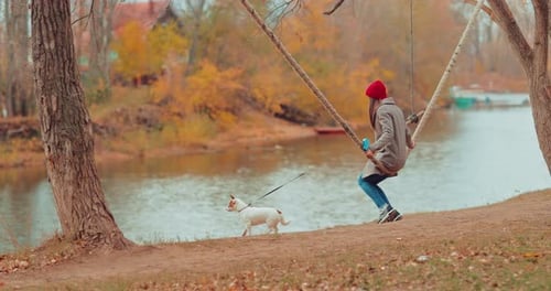 Young Woman is Swinging on a River Swing and Holding a Puppy on a Leash