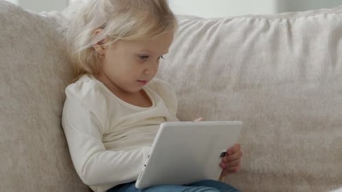 Little Girl Using a Digital Tablet While Sitting on the Couch Watching Cartoons or Browsing the