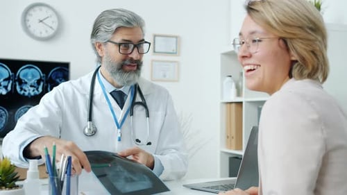 Happy Young Woman Getting Good News From Doctor Holding Hands Looking at MRI Scans in Clinic