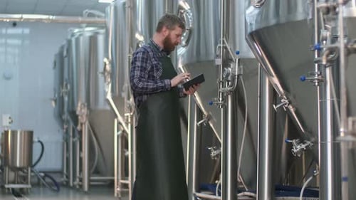 A Male Brewer with a Beard Monitors the Readings on Beer Tanks Using a Tablet. Craft Beer Production