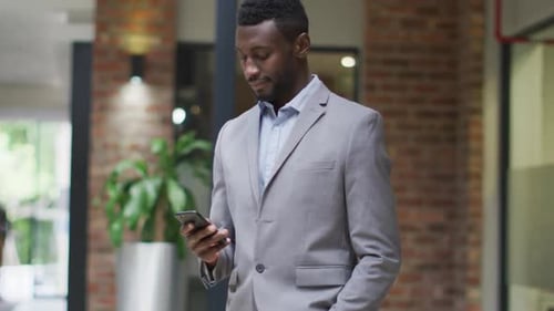 Smiling african american businessman standing in corridor using smartphone in modern office