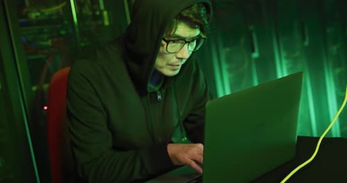 Asiatischer männlicher Hacker im Hoodie benutzt Laptop von Computerservern