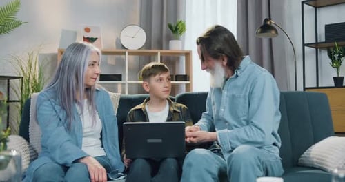 Abuelo sentado en el sofá con su nieto adolescente que estudia entre ellos en una computadora portátil