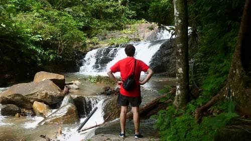 Traveler Man in Red TShirt Standing on Stone Near Waterfall in Tropical Forest