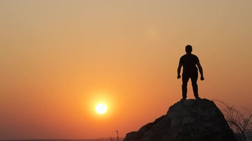 Silhouette of a man hiker jumping alone on big stone at sunset in mountains. Male tourist raising