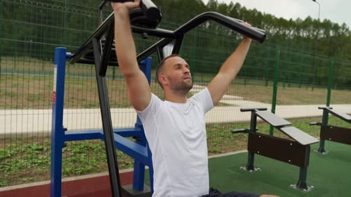 Young Man Trains Outdoors on a Sunny Summer Day Trains His Shoulders on a Simulator in the Park