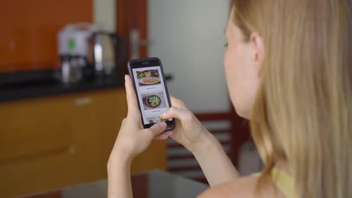 Young Woman Orders Food Online Using a Smartphone. She Is Sitting on in Her Kitchen in Her Apartment