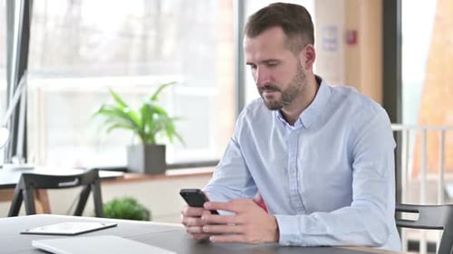 Smartphone Use By Young Man with Laptop in Office