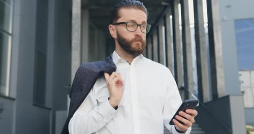Office Manager in white Shirt which Walking Near Modern Office Building and Uses His Smartphone