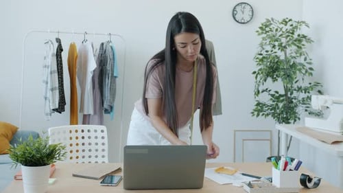 Selbstbewusste asiatische Näherin, die drinnen im Studio mit dem Laptop tippt und Material auswählt