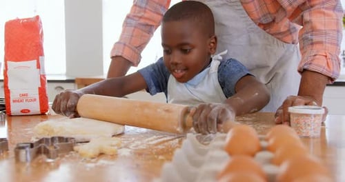 Front view of little black son rolling dough with rolling pin in kitchen of comfortable home 4k