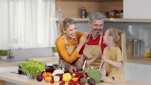 Happy Parents and Little Daughter Preparing Dinner Together at Kitchen Mixing Vegetable and Laughing