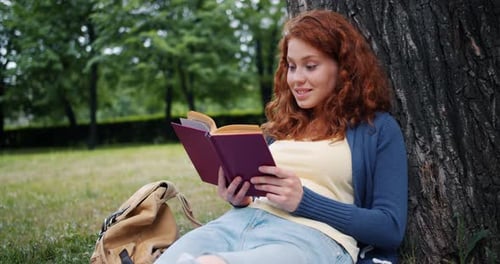 Cheerful Redhead Girl Reading Book Outdoors in Park Smiling Relaxing on Grass