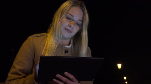 A Young Beautiful Woman Sits on a Bench and Works on a Tablet in an Urban Area at Night