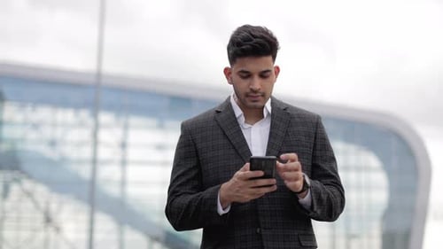 Indian Businessman in Formal Jacket Standing Near Modern Building and Texting on Smartphone