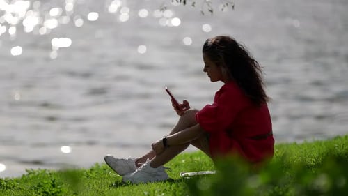 Relaxed Young Woman is Spending Time on Shore of River or Lake and Using Smartphone