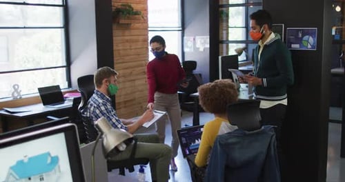 Diverse colleagues wearing face masks with electronic devices discussing together at modern office