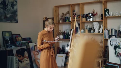 Girl Painter Is Working in Workroom Alone Holding Palette and Brush and Looking at Canvas