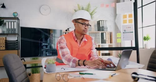 Ingeniero masculino con casco y chaleco trabajando con un proyecto de computadora y un plan en papel