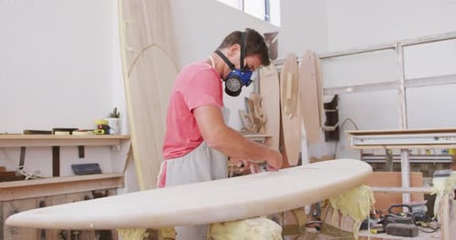 Caucasian male surfboard maker wearing a face mask and preparing to polishing a surfboard