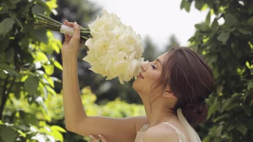 Beautiful Stylish Bride in White Wedding Dress and Veil Holding Wedding Bouquet in Hands in Park