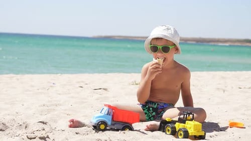 Tanned Boy Sits on a Sandy Beach and Eats Ice Cream