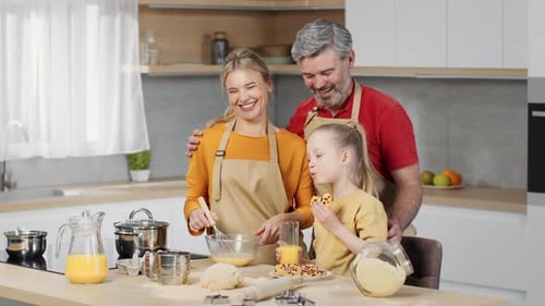 Cute Little Girl Eating Cookie and Drinking Juice at Kitchen Her Mother and Father Cooking Near at