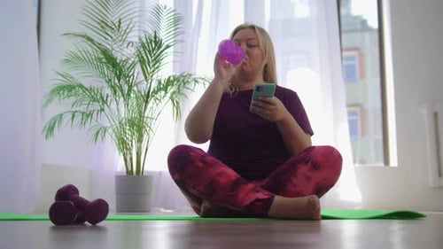 Woman Drinks Water and Checks Phone on Yoga Mat