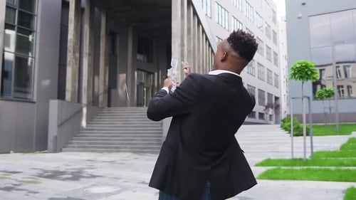 Black-Skinned Man Dancing in the Street Near Modern Building and Scattering One Hundred Dollars