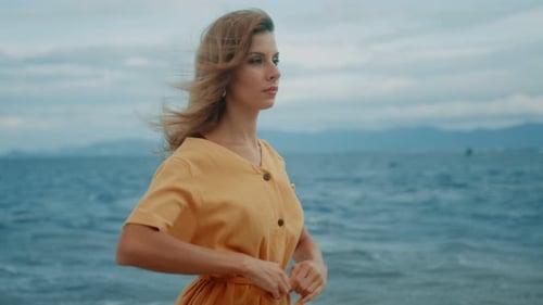 A Slim Blonde in a Yellow Dress Walks Along the Beach and Looks Around