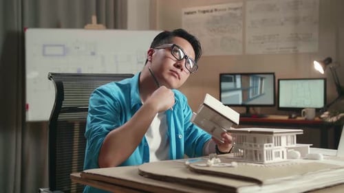 Asian Man Engineer With A Laptop Thinking While Holding Paper Model Of House At The Office