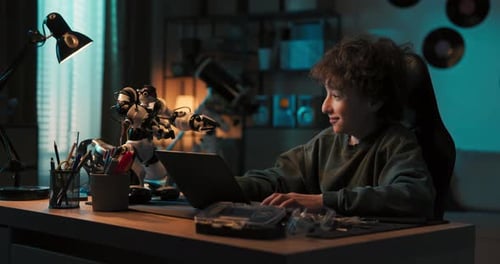 Young Talented Gifted Boy Sits in Room in the Dark and Programs a Robot on Laptop Adds New Features