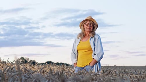 Image Of Blonde In Glasses And Hat. Woman Stands On Wheat Field And Smiles.