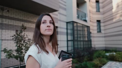 Woman Carries Tablet and Coffee Outside Apartment Building