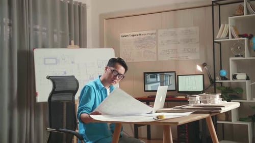 Asian Man Engineer Typing On A Laptop And Looking At Blueprint Before Walking Out Of The Office